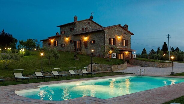 Una casa de piedra con piscina, mobiliario de exterior y un jardín bien iluminado al anochecer.