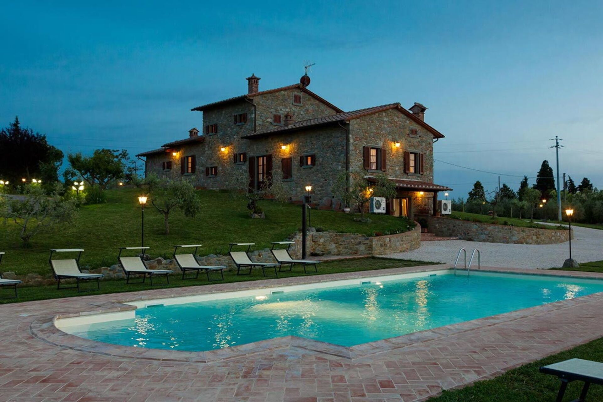A stone house with a swimming pool, outdoor seating, and a well-lit garden at dusk.
