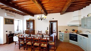 Un comedor con una mesa de madera dispuesta para seis personas, rodeada de sillas. Sobre la mesa hay una araña, y a la derecha se ve una zona de cocina con armarios y electrodomésticos.