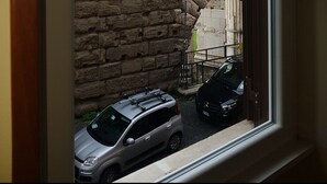 A view through a window of a stone structure with columns, a parked car, and a brick wall. - Residenza Maritti Classic Rooms (Rome)