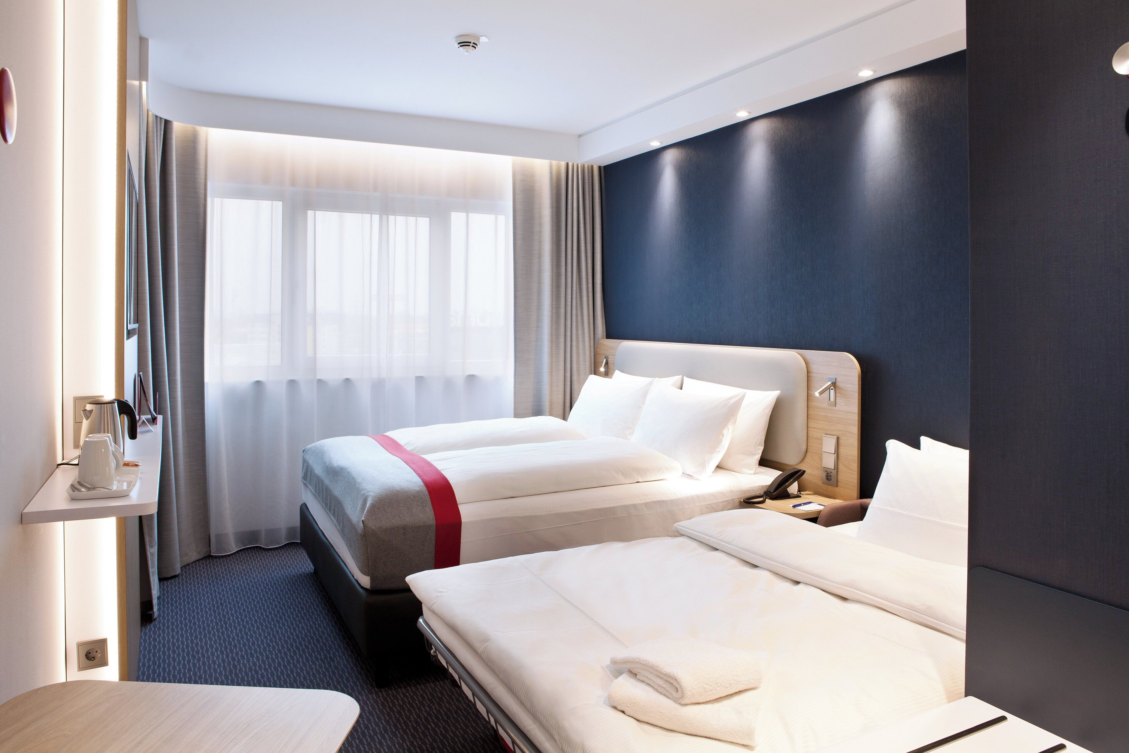 A hotel room with two beds, a bedside table, a desk, and a window with curtains.