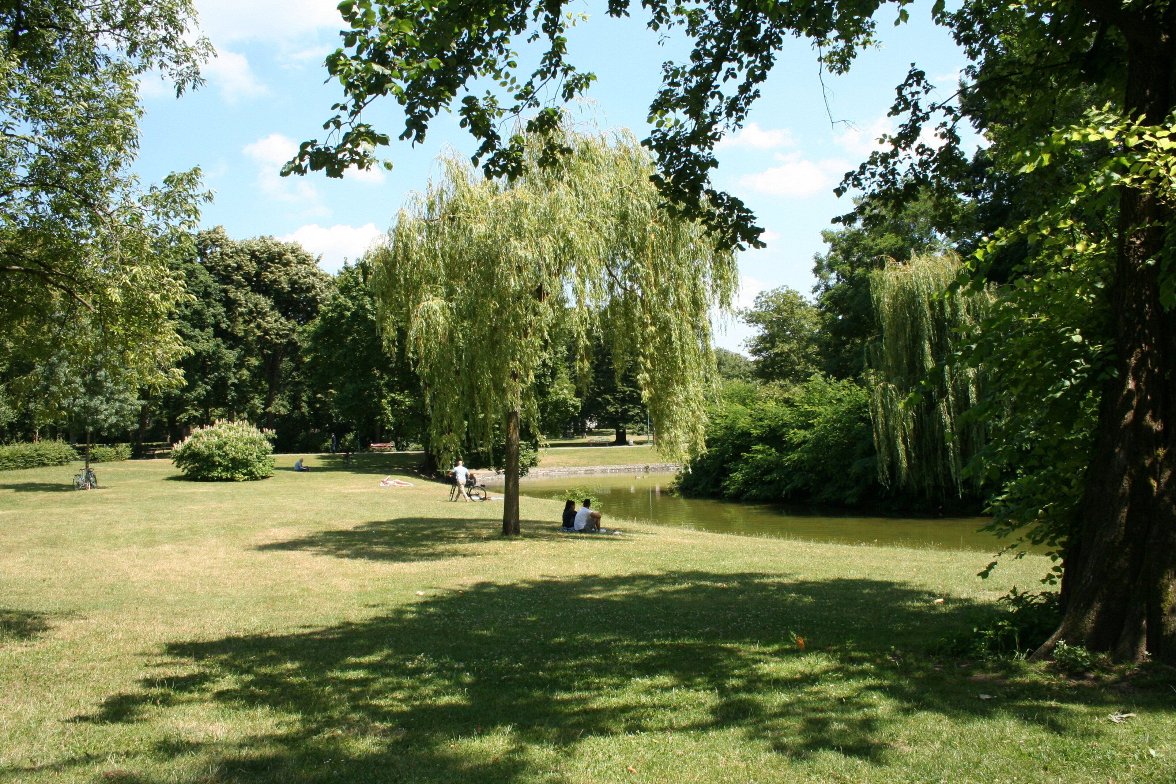 A park with a pond, trees, and people sitting on the grass.