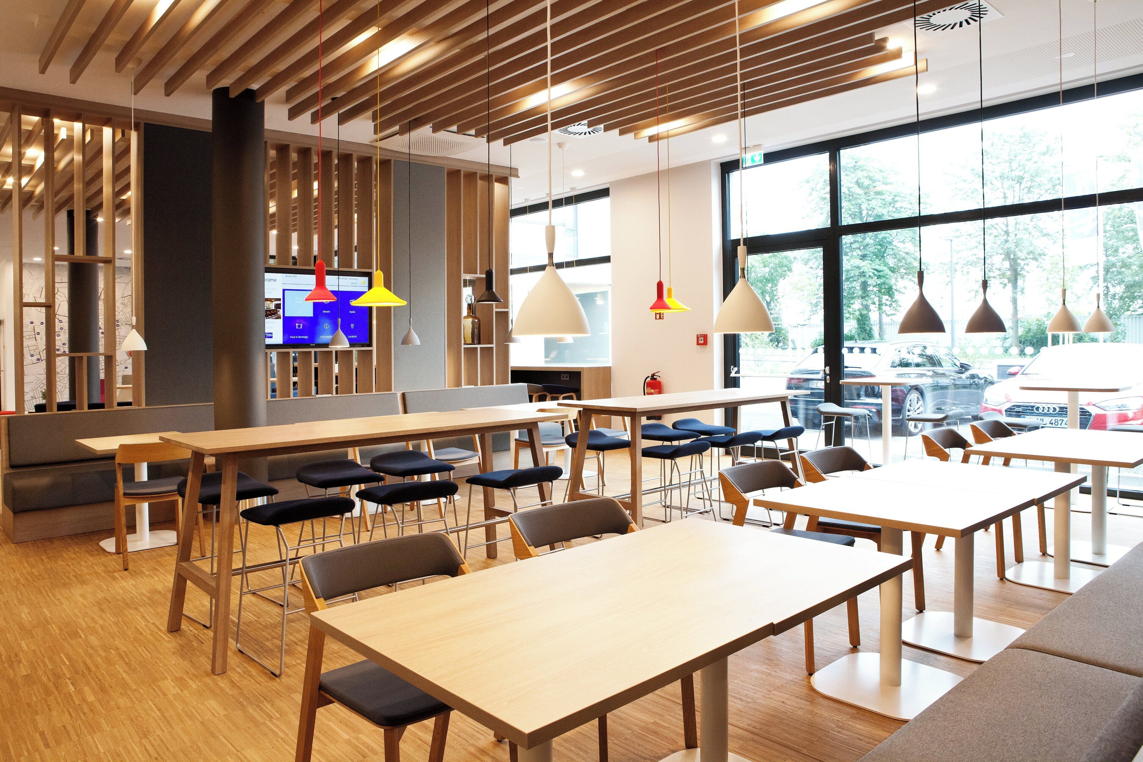 A modern restaurant with wooden tables and chairs, a digital menu display, and large windows offering a view of the outside.