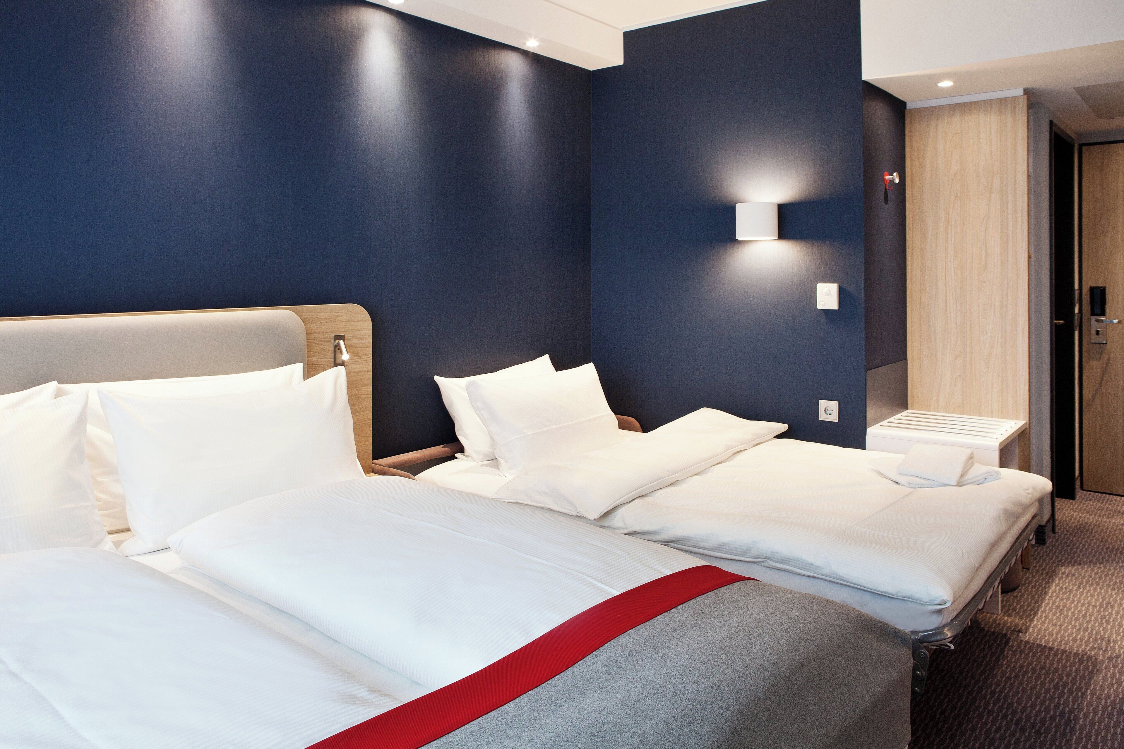 A hotel room with two beds, a wooden headboard, a bedside table, and a wall-mounted light.