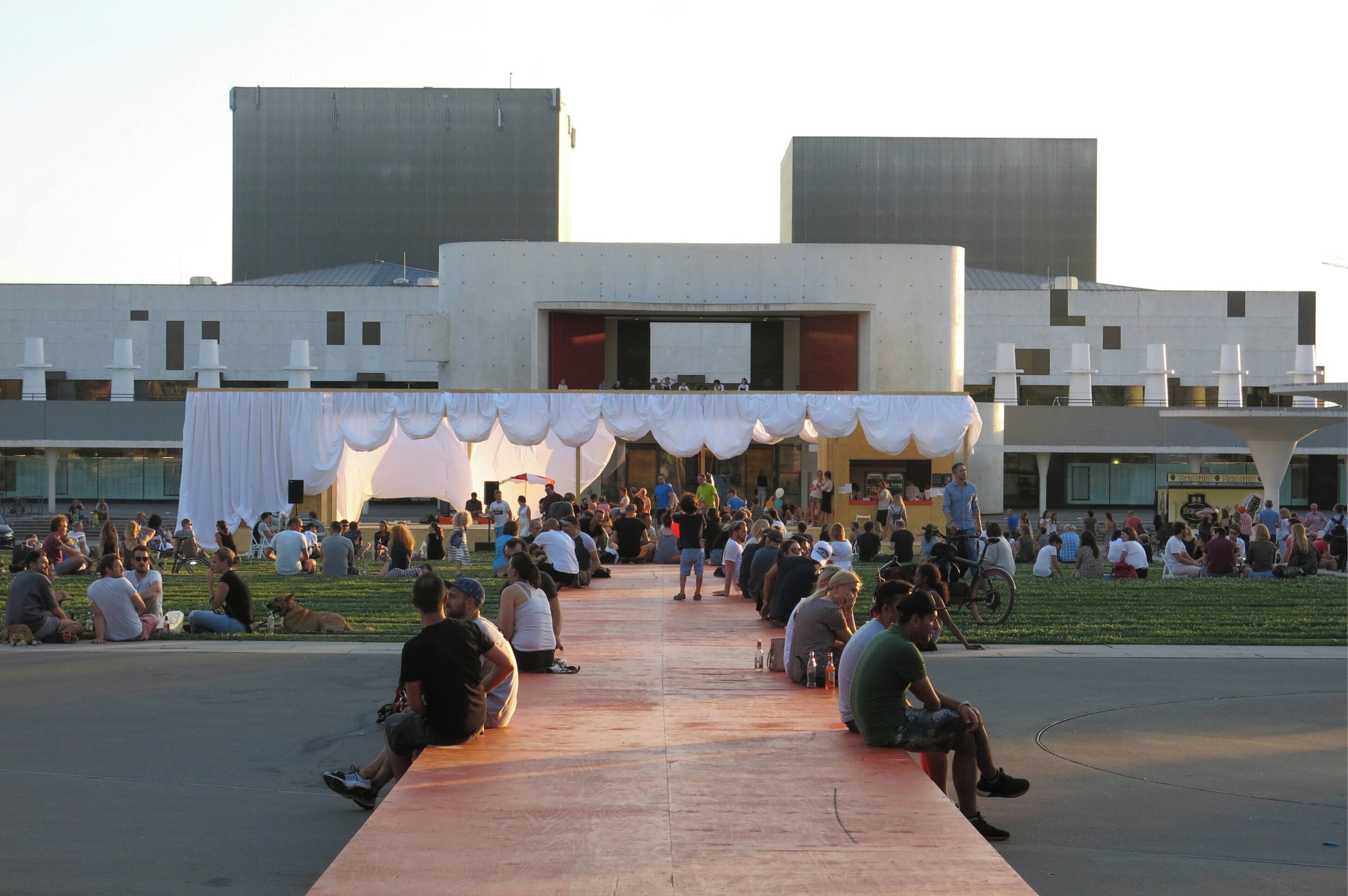 Una plaza pública con un edificio moderno, un espacio abierto central y una zona cubierta.
