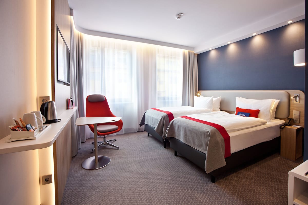 A hotel room with two beds, a desk, a chair, and a large window with curtains.