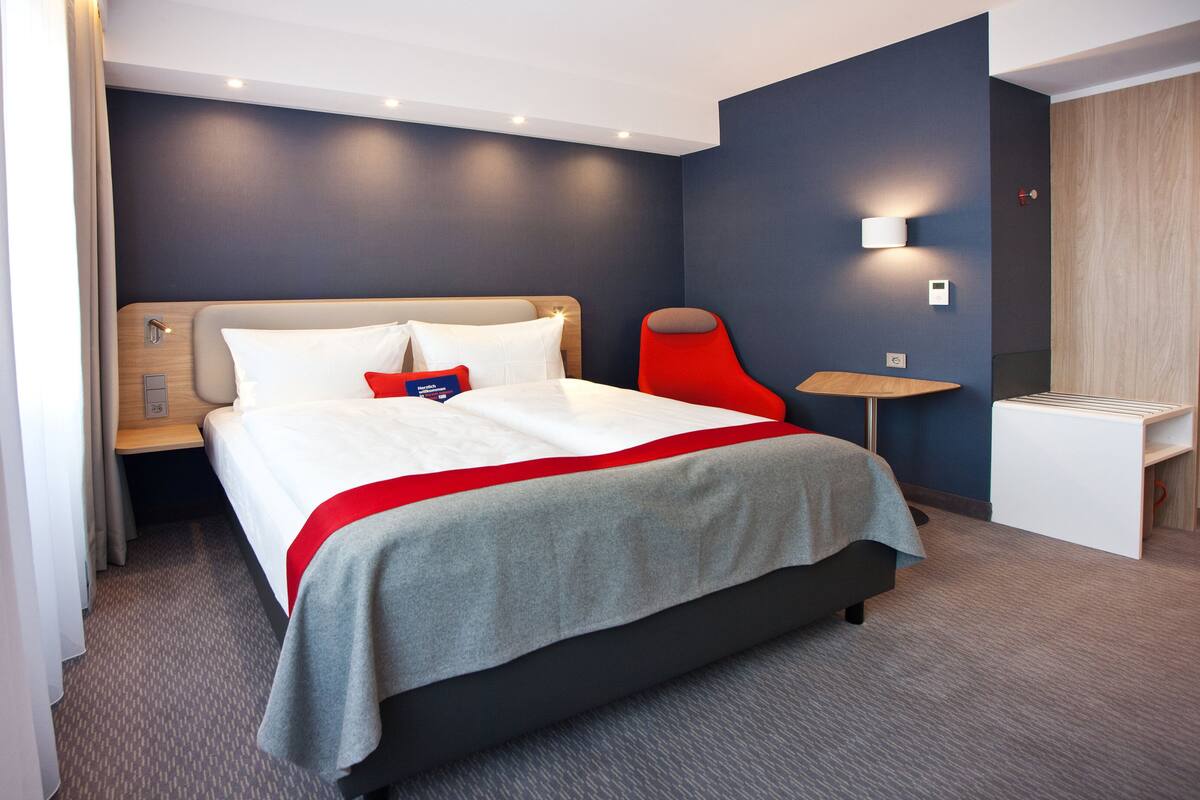 A modern hotel room with a large bed, a bedside table, a small desk, and a wall-mounted lamp.