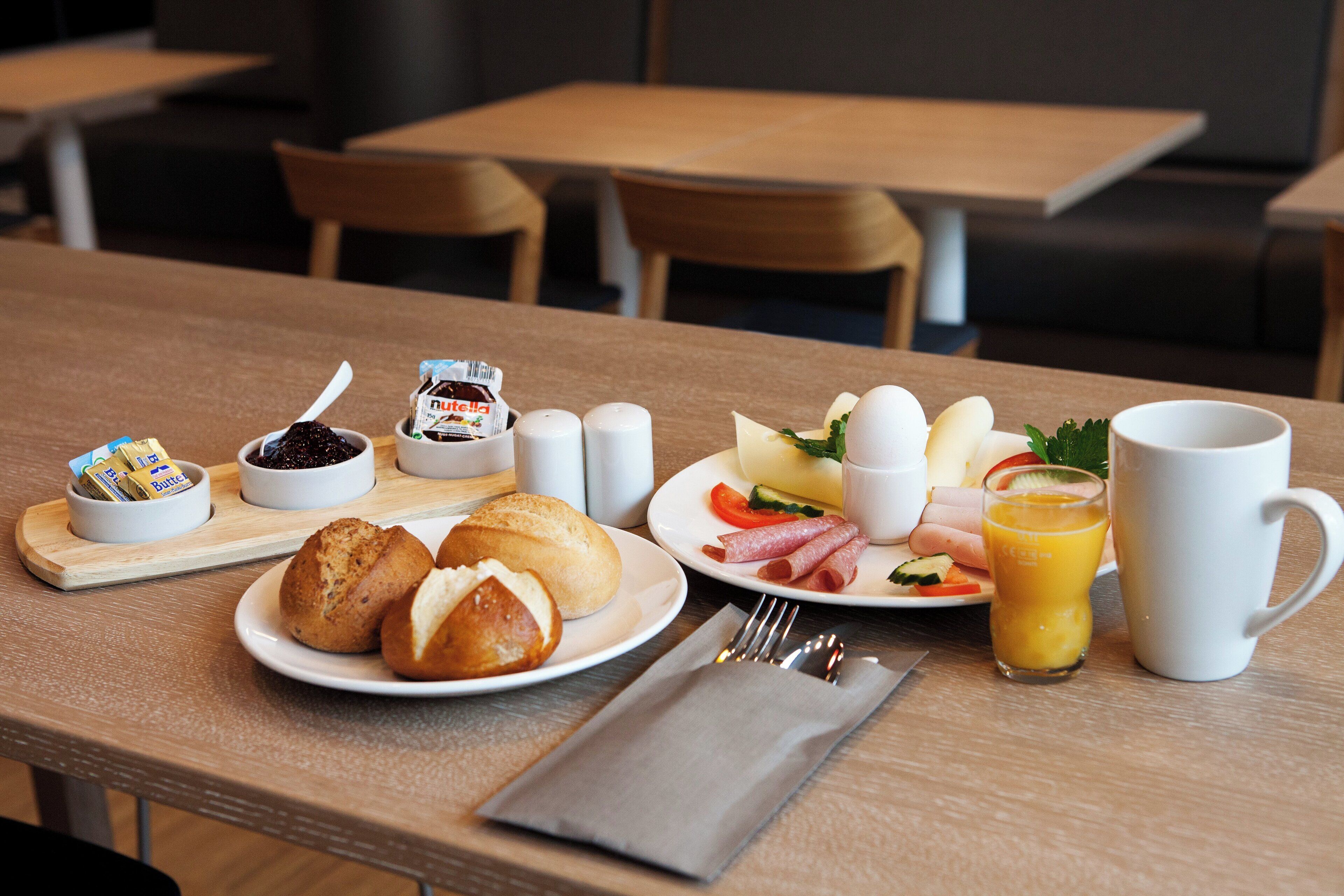 A breakfast table with bread, butter, jam, eggs, ham, vegetables, and a glass of orange juice.