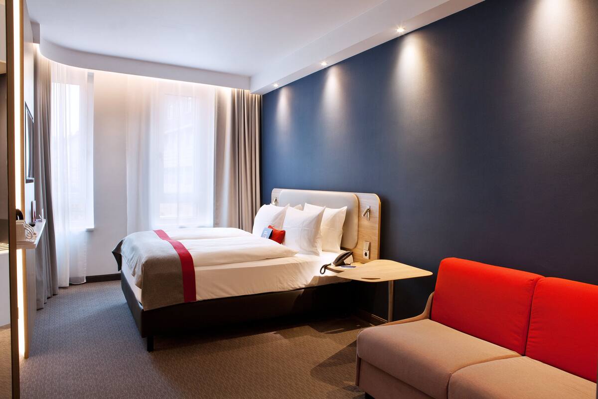 A modern hotel room with a large bed, a red sofa, and a desk with a telephone.