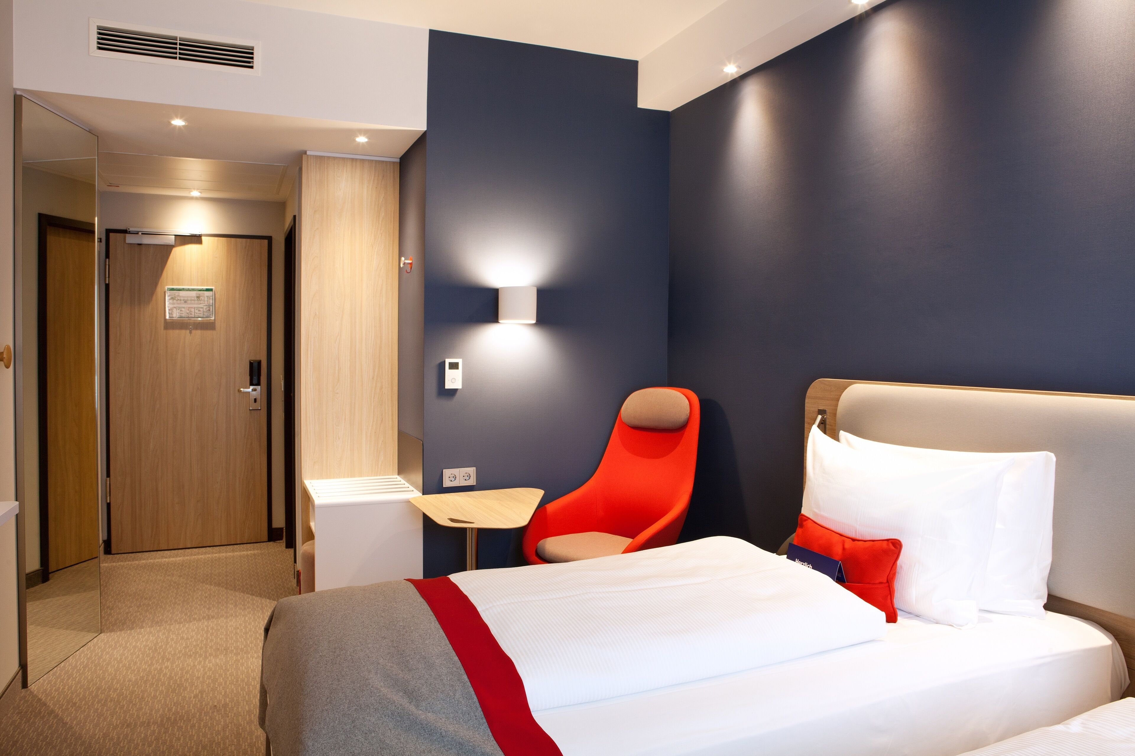 A hotel room with a bed, a red chair, a small table, and a wall-mounted light.