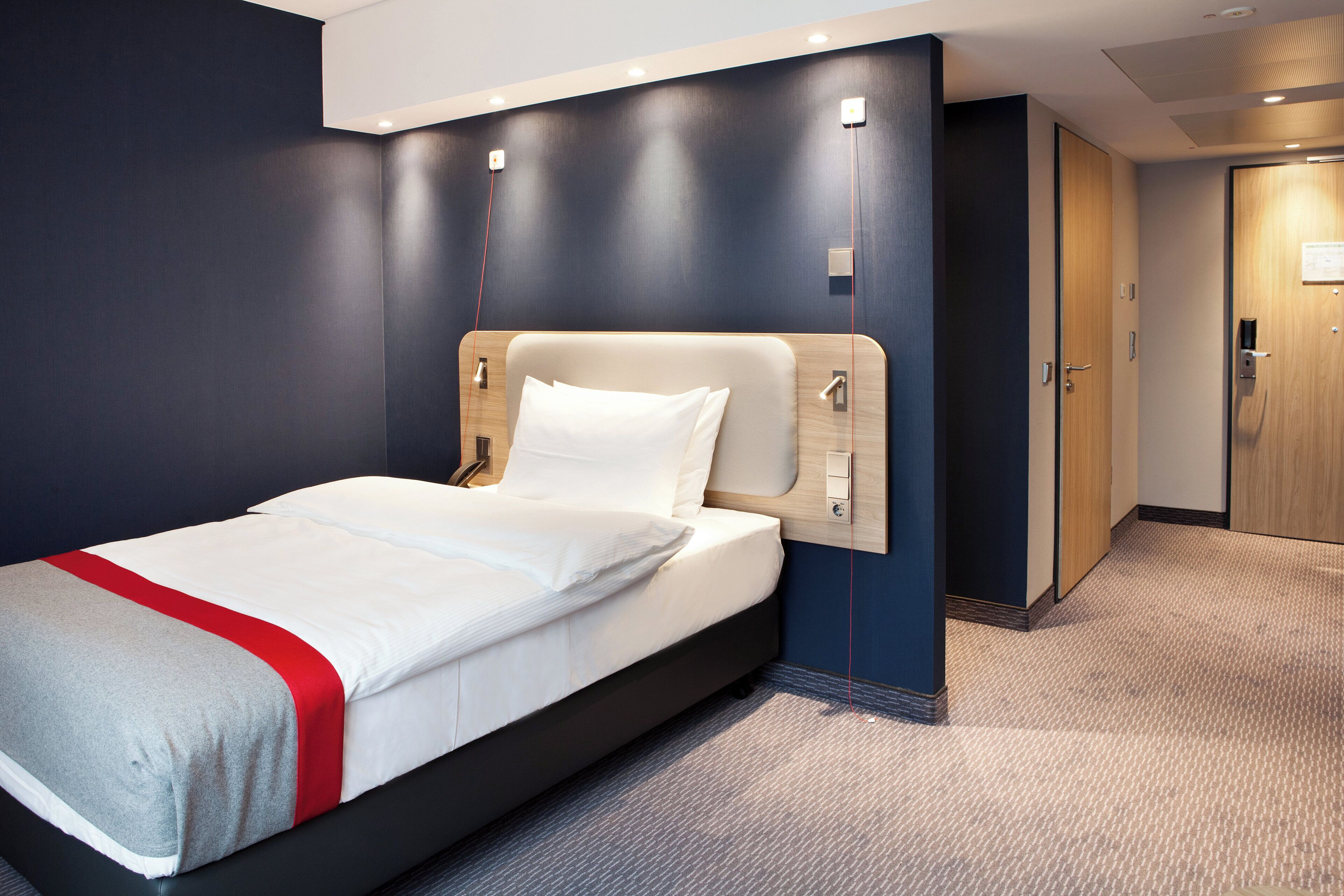 Habitación de hotel con cama, cabecero de madera, un detalle rojo en la colcha, un panel de control en la pared y una puerta a la derecha.
