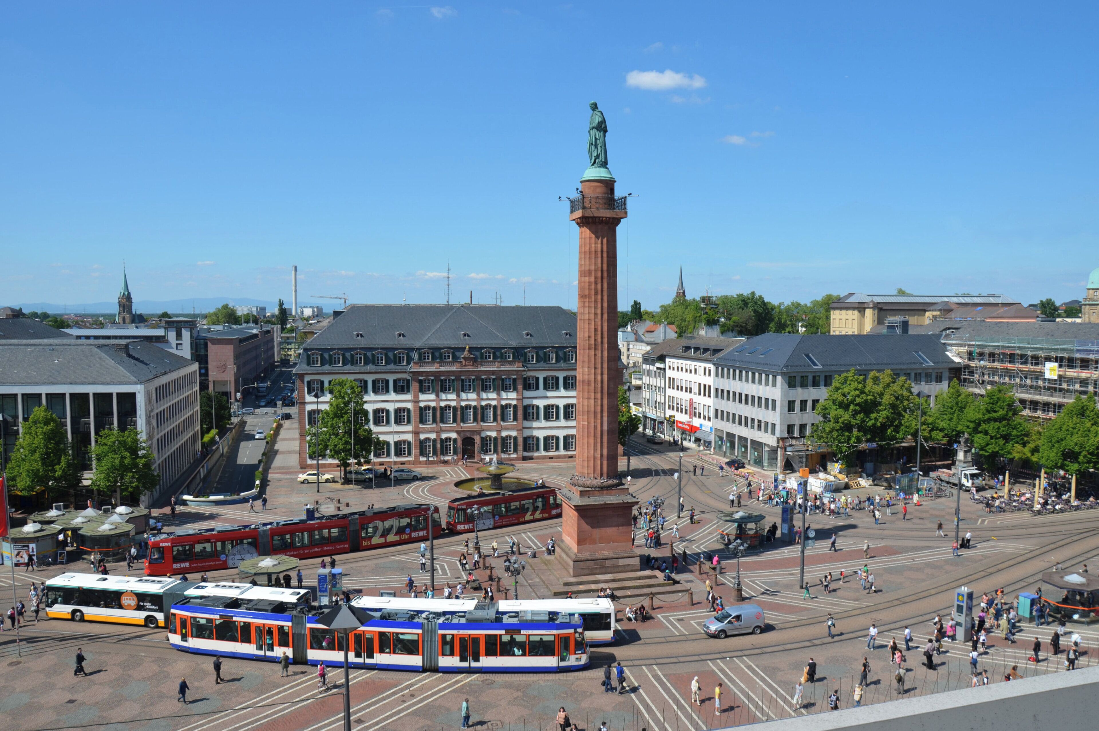 A city square with a prominent red monument, tram lines, and surrounding buildings.