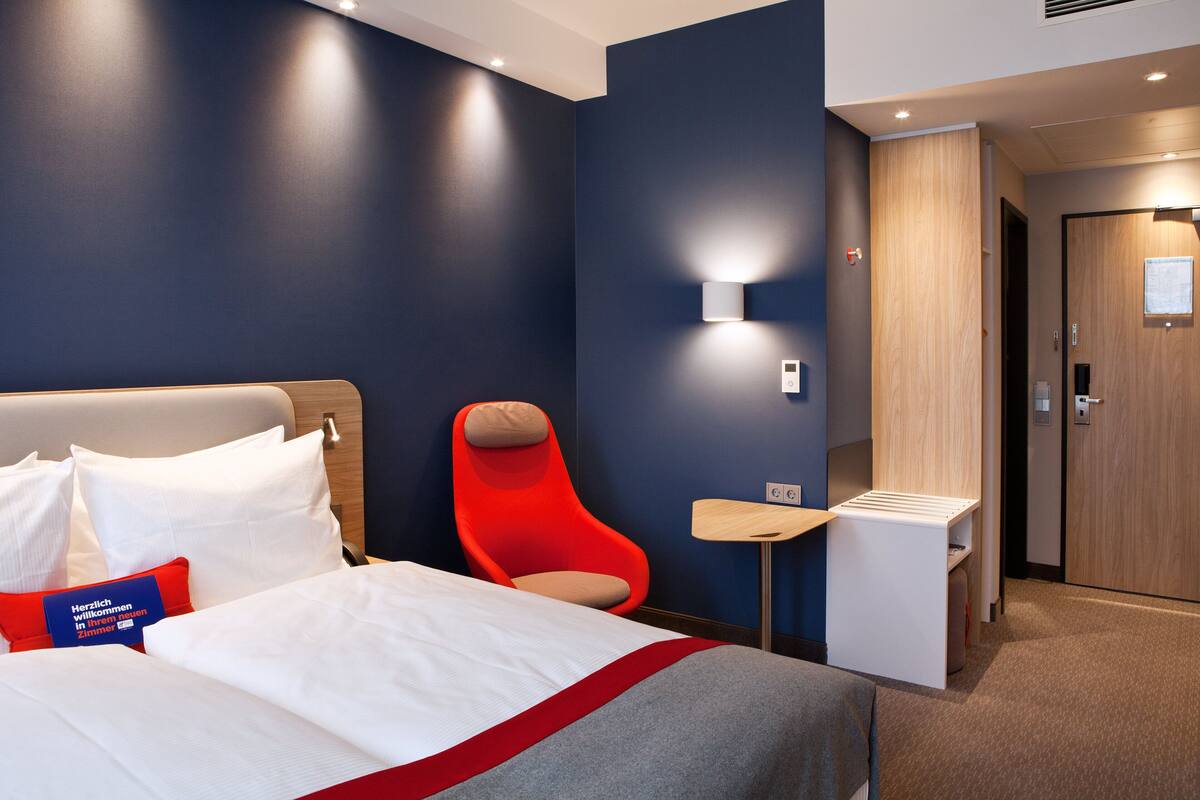 A modern hotel room with a large bed, a red chair, a small table, and a wardrobe.