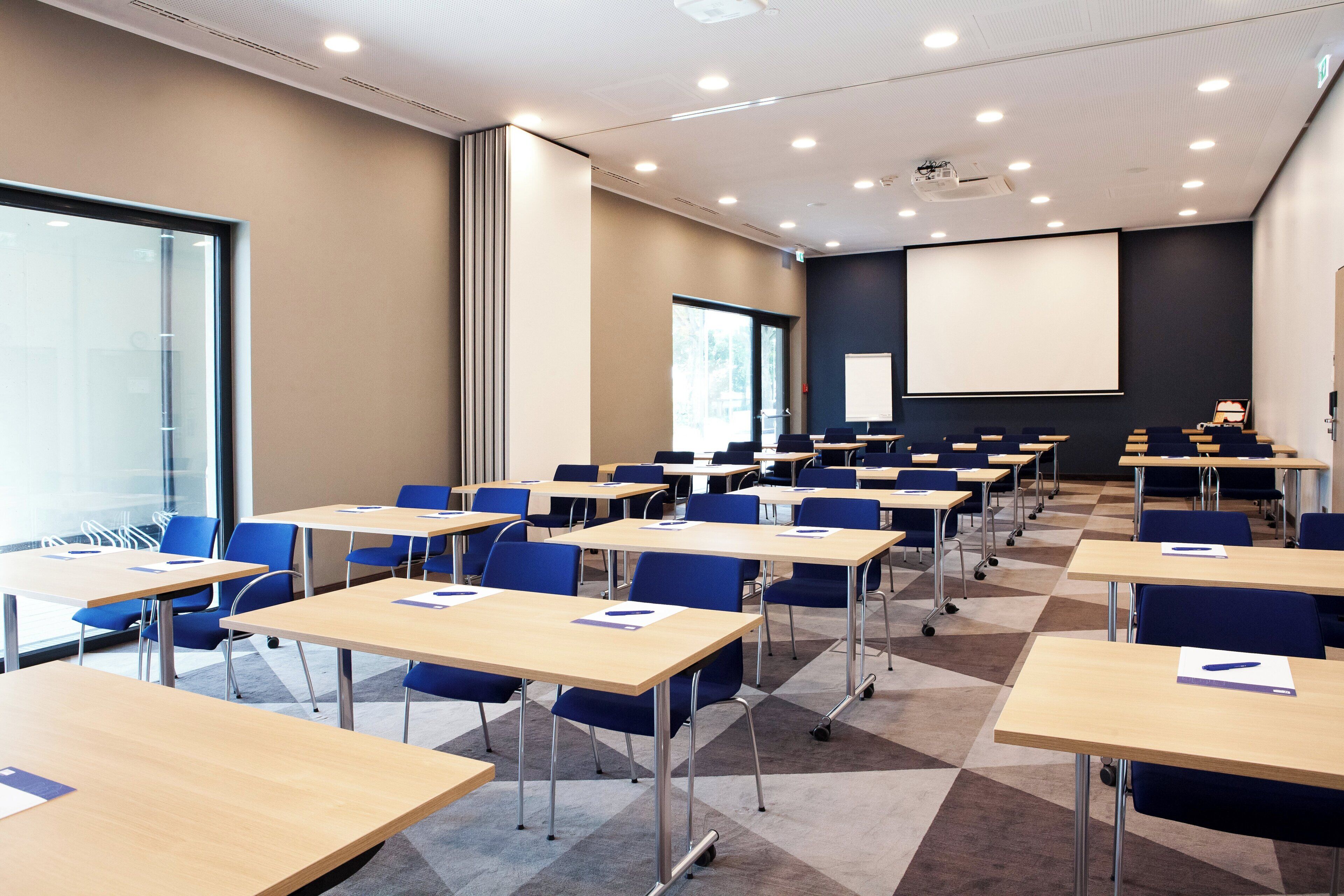 Uma sala de conferências com várias mesas e cadeiras, uma tela grande e um projetor.