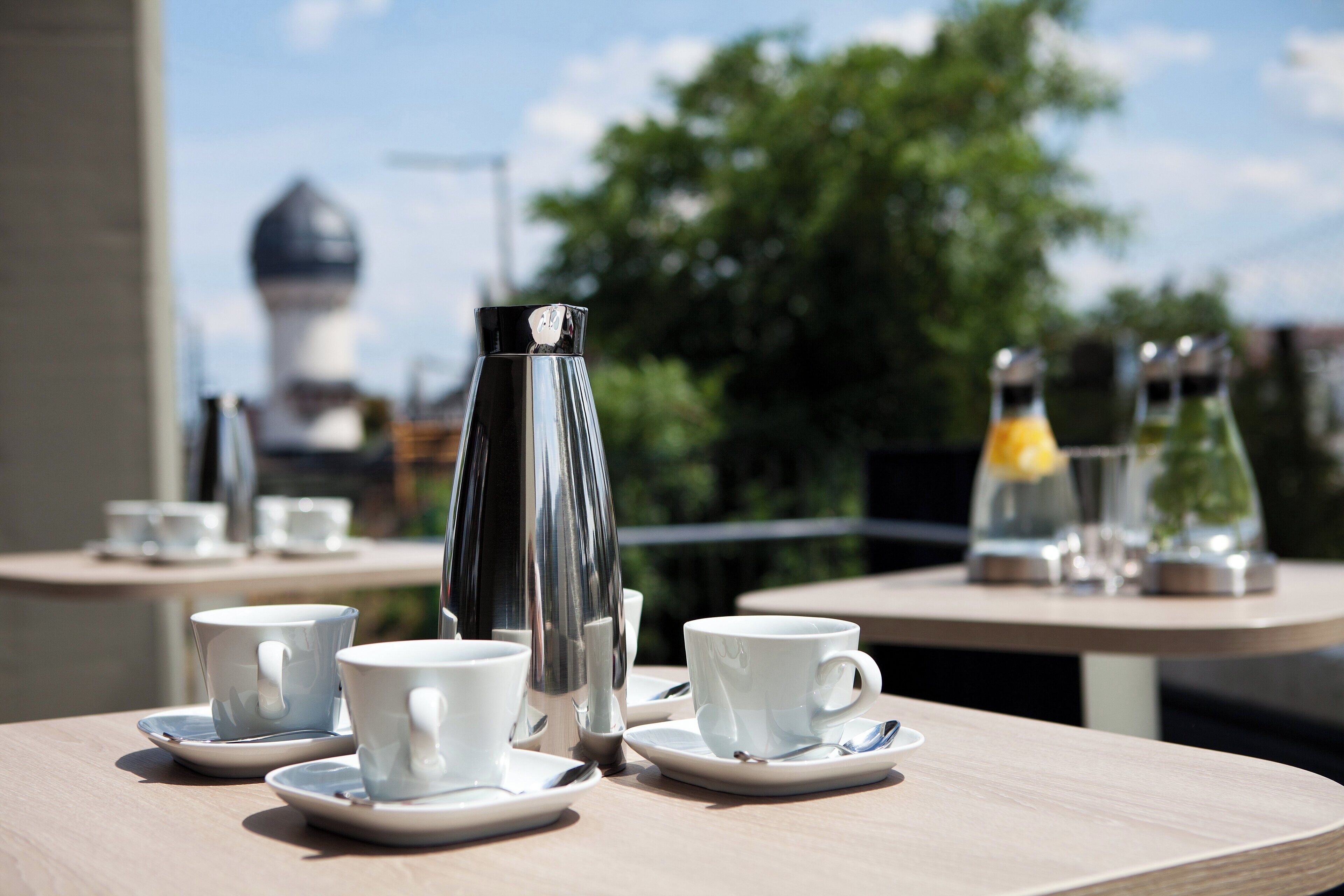 A table with white cups and saucers, a silver pitcher, and a glass bottle, set outdoors with a view of trees and a building in the background.
