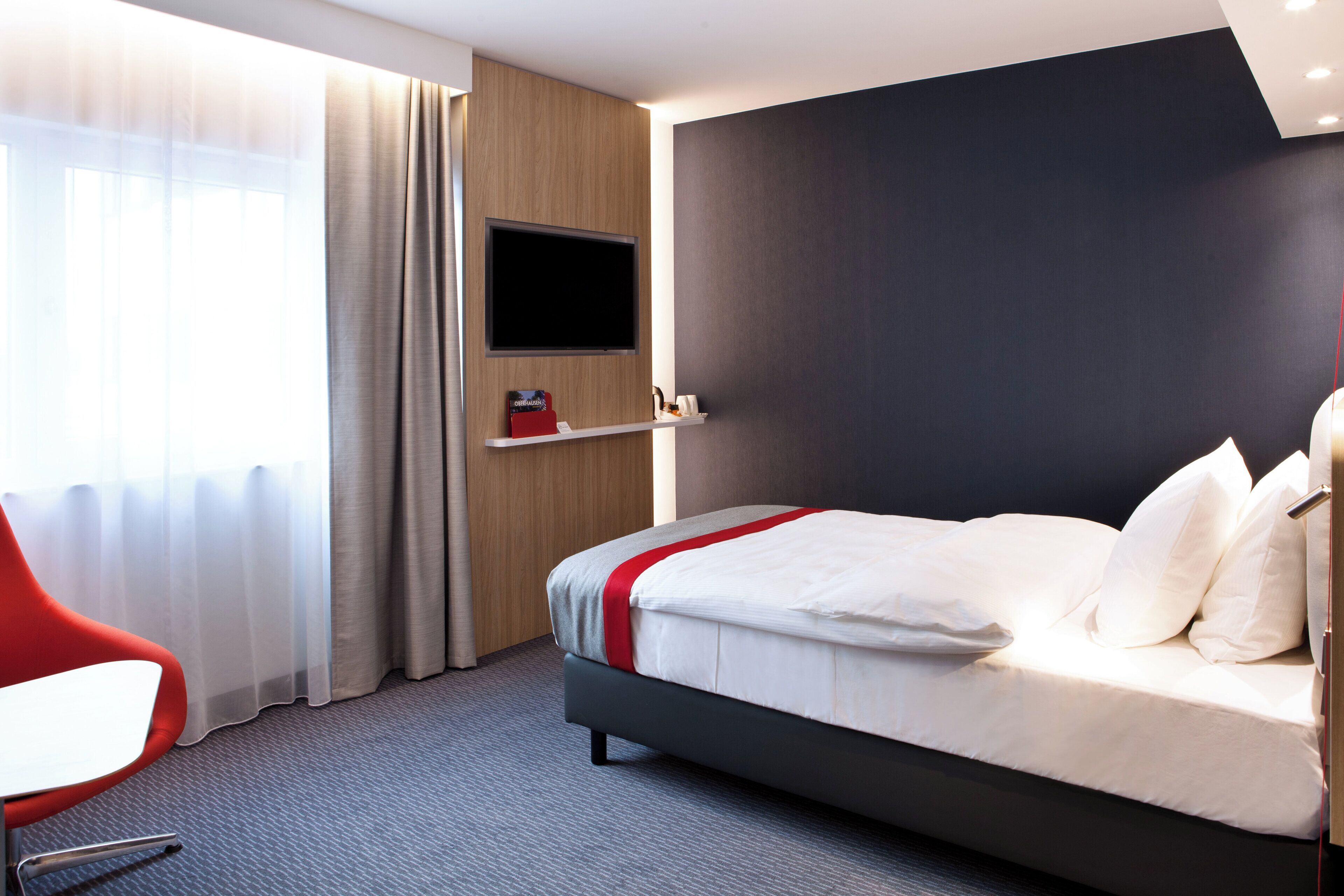 A hotel room with a bed, a TV mounted on the wall, a red chair, and a window with curtains.