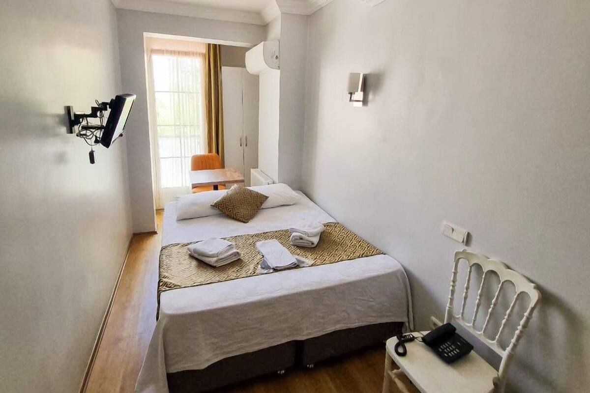 A hotel room with a bed, a chair, a desk, a wall-mounted TV, and a window with curtains.