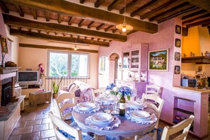 Zona pranzo con tavolo rotondo apparecchiato per sei persone, circondato da sedie. Alla sinistra, una televisione su un mobile, e un camino con mensola. La stanza presenta travi a vista al soffitto e una finestra con vista sugli alberi.