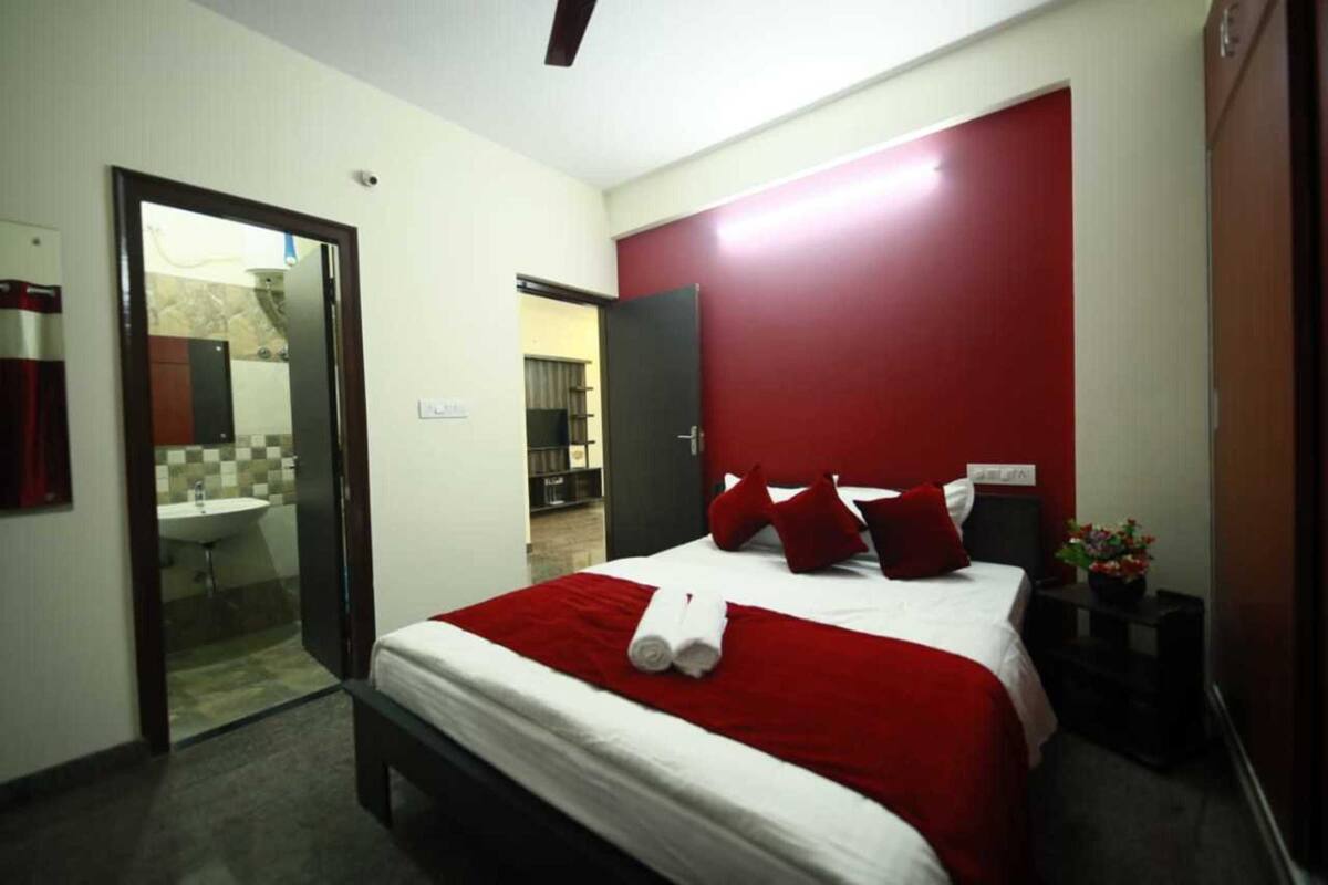 Una habitación de hotel moderna con una cama grande, ropa de cama roja y blanca, una mesita de noche con una planta y una pared de acento roja.