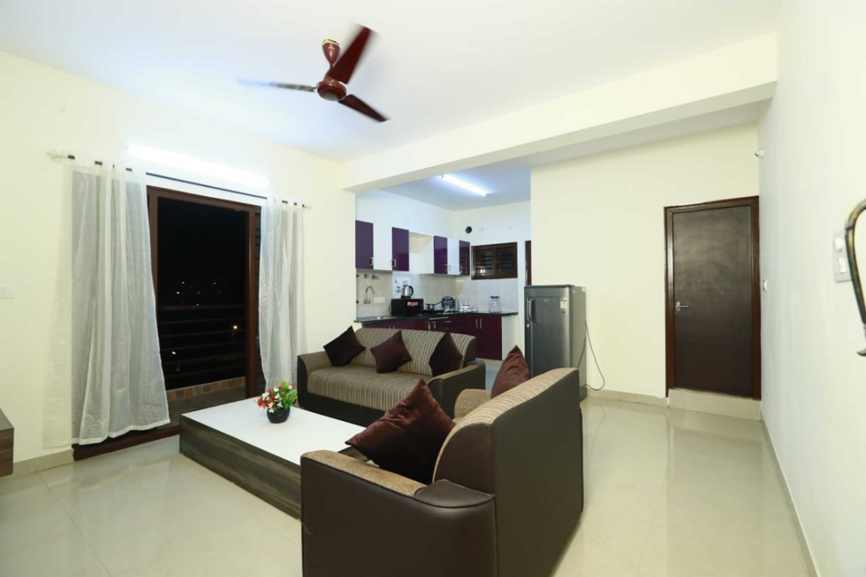A living room with a sofa, a coffee table, and a ceiling fan. There is a kitchen visible through an open doorway.