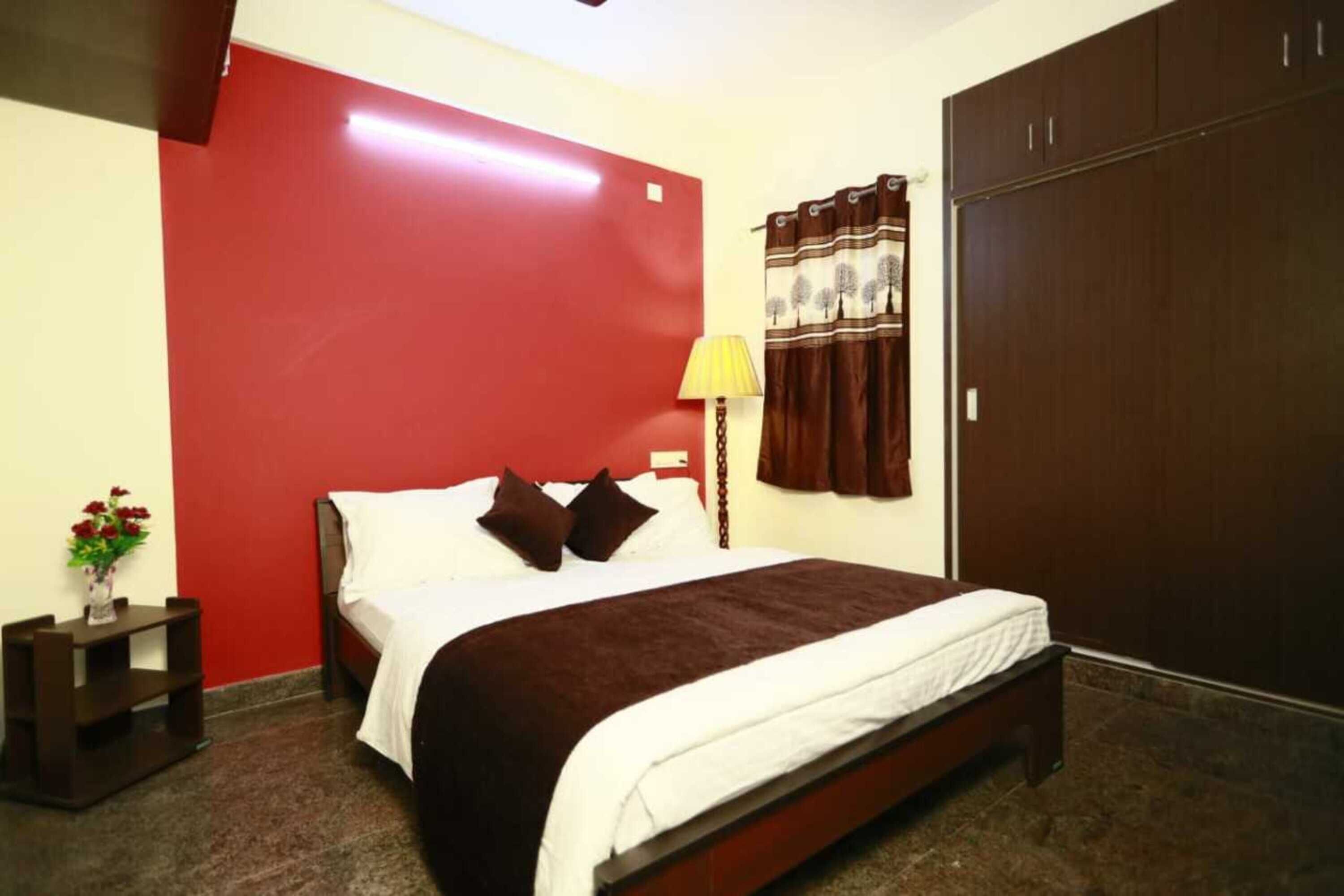 A bedroom with a bed, a nightstand, a lamp, a wardrobe, and a red accent wall.