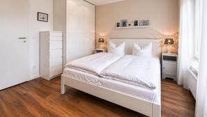Un dormitorio con cama, armario, mesita de noche con lámpara y estante de pared con fotografías.
