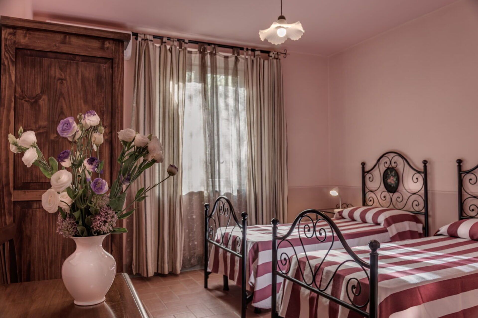 A bedroom with two beds, a wooden door, a window with curtains, and a vase with flowers on a table.
