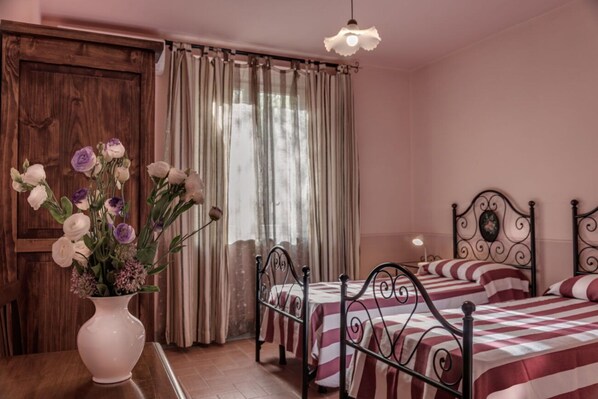 A bedroom with two beds, a wooden door, a window with curtains, and a vase with flowers on a table.