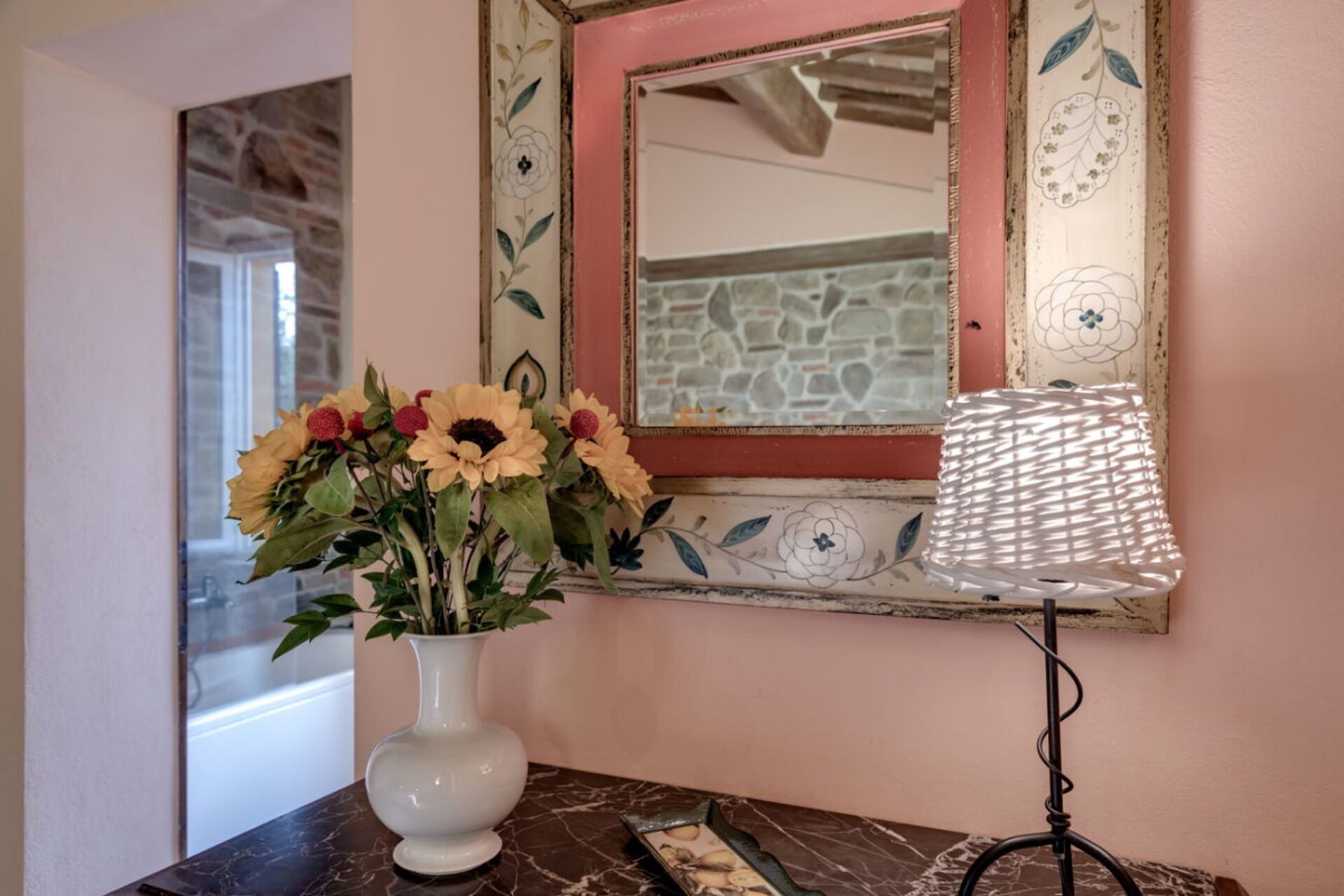 Un vase blanc orné de tournesols, posé sur une surface en marbre, un miroir décoratif aux motifs floraux et une lampe avec un abat-jour blanc.