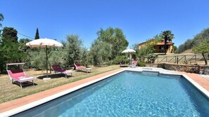 A rectangular swimming pool with clear blue water, surrounded by a stone border. There are lounge chairs with umbrellas and a small poolside area.