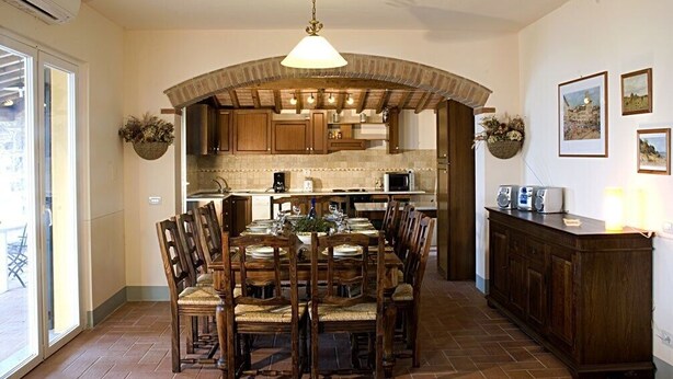 Un espace repas avec une grande table et des chaises en bois, un luminaire suspendu et une cuisine en arrière-plan.