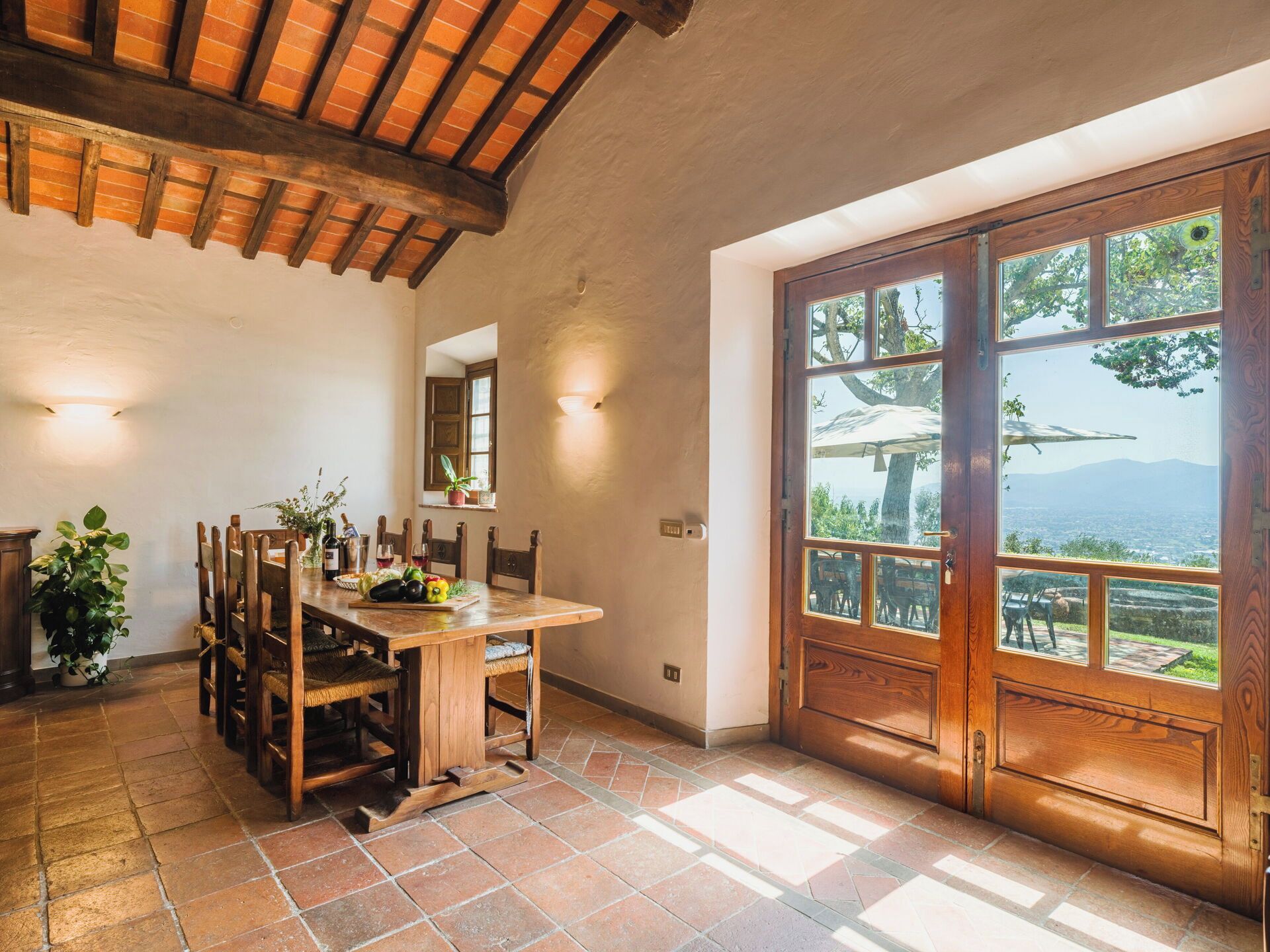 A dining area with a wooden table and chairs, a potted plant, and a view through a large wooden door.