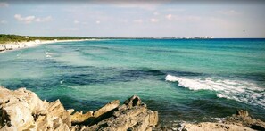 Een kuststrand met helder turquoise water, een rotsachtige kustlijn en gebouwen in de verte.