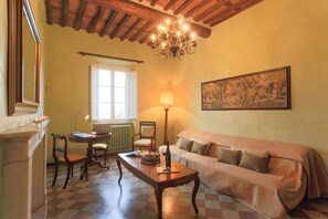 Une pièce chaleureuse avec un plafond en bois, un lustre, un canapé, une table avec un ordinateur portable, une chaise et une fenêtre avec des volets.