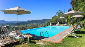 Une piscine entourée d’une terrasse en pierre et d’une végétation luxuriante, avec des chaises longues et des parasols à proximité.