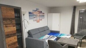 Ein modernes Wohnzimmer mit einem grauen Sofa, einem Couchtisch mit einem blau-weißen Tischläufer, einem grauen Sessel und einem weißen Kleiderschrank.