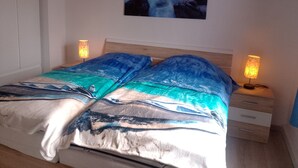 Ein Schlafzimmer mit einem Bett, das mit einer blauen und weißen Bettdecke im Meeresstil, zwei Nachttischlampen, einem Nachttisch und einem Wandbild einer mondbeschienenen Meereslandschaft ausgestattet ist.