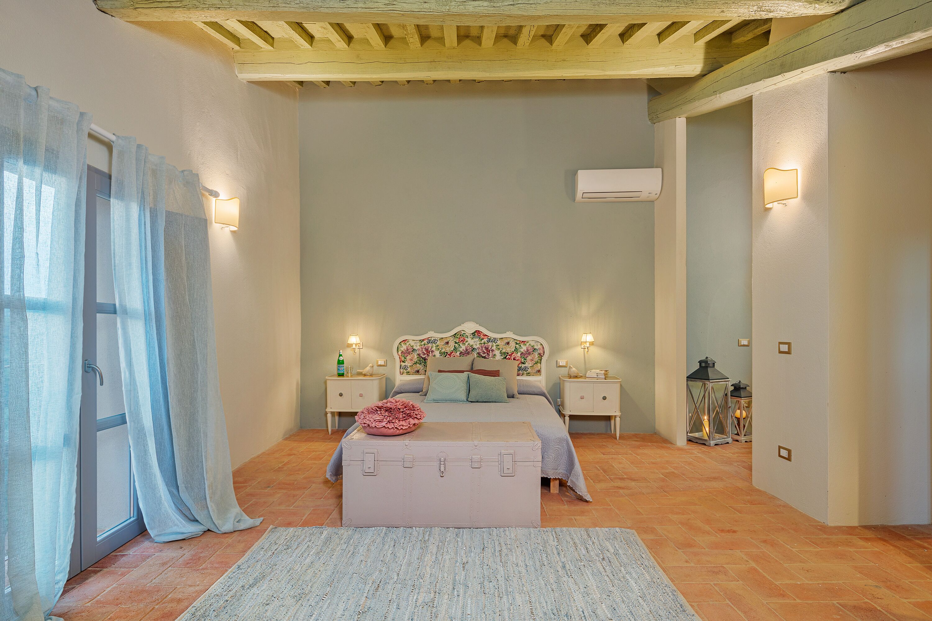 Un dormitorio con cama, lámparas de noche, una maleta y una mesita de noche con una botella.