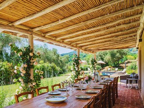 Ein Essbereich im Freien mit einem langen Holztisch, gedeckt für eine Mahlzeit, umgeben von Stühlen, mit Blick auf einen Pool und viel Grün im Hintergrund.