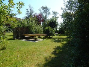 A grassy outdoor area with a wooden bench and a small brick structure, surrounded by trees and shrubs.