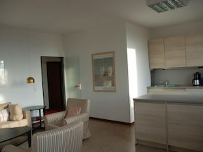 A modern living room with a sofa, armchair, and a small table. A kitchen area with cabinets and a sink is visible.