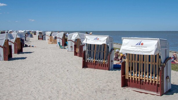 Beach huts with white canopies, arranged in rows along a sandy beach, with the sea in the background.