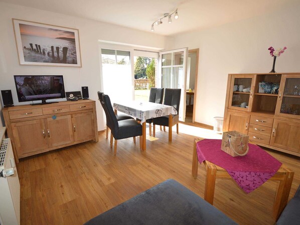 A cozy living room with a wooden floor, a dining table with chairs, a television, and a cabinet with glass doors.
