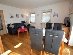A modern living room with a sofa, dining table, and chairs. There are framed pictures on the walls and a window with curtains.