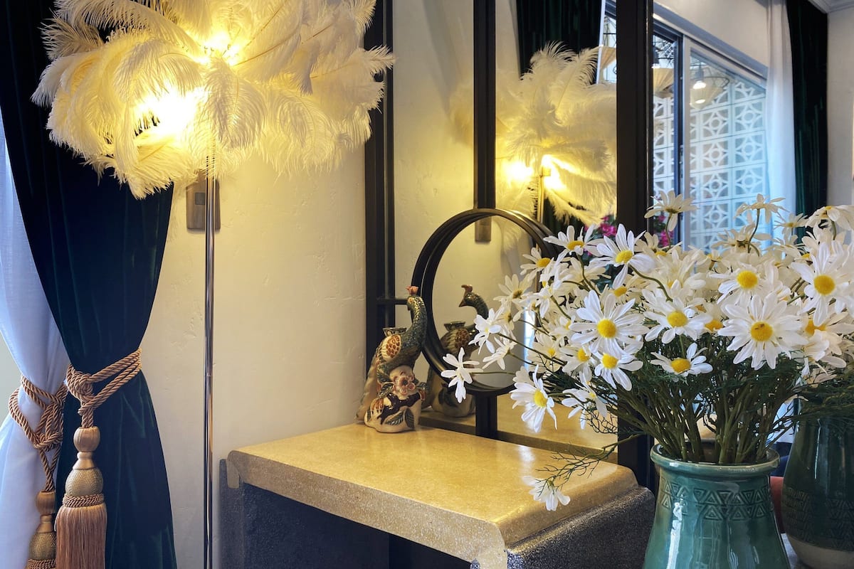 Een kamer met een spiegel die een decoratieve haan, een vaas met witte bloemen en een verenlampatuur weerspiegelt.