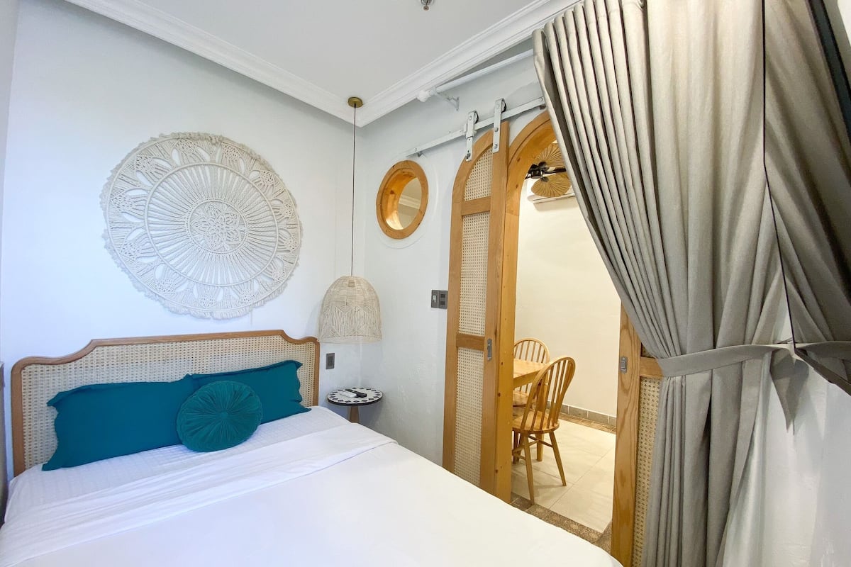 Een slaapkamer met een bed, een ronde wanddecoratie, een spiegel met houten lijst en een eethoek met stoelen.