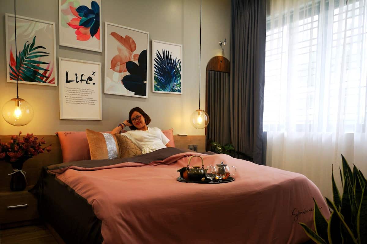 Een keurig opgemaakt bed met een roze dekbed, een nachtkastje met een bloemenvaas, en lijstwerk op de muur.
