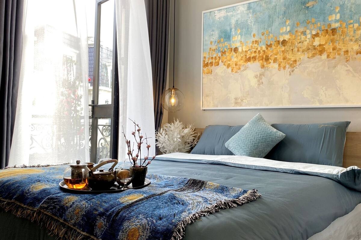 Een moderne slaapkamer met een groot bed, een stadsgezicht schilderij en uitzicht op gebouwen door het raam.