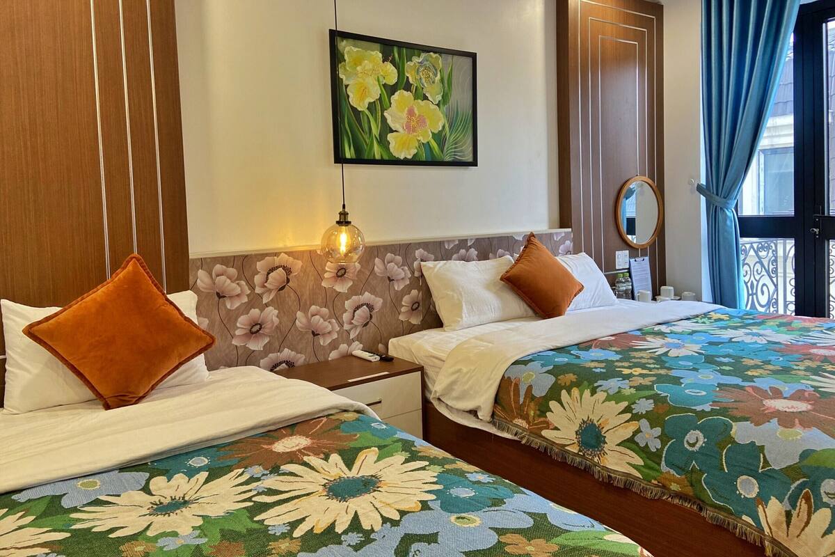 Een hotelkamer met twee bedden, hoofdborden met bloemenprint, een aan de muur hangend schilderij, een nachtkastje met een lamp en een raam met gordijnen.