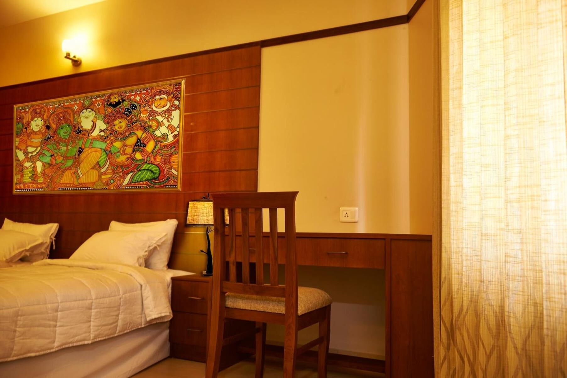 A hotel room with a bed, a desk, and a chair. There is a large colorful tapestry on the wall above the bed.
